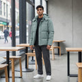 City Center sage green hooded puffer jacket worn by a man with dark trousers and white sneakers in a modern cafe