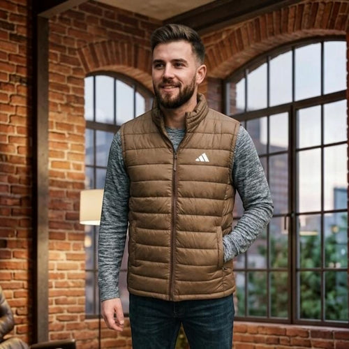City Center Adidas brown quilted puffer vest worn by a man over a grey long-sleeve shirt in a brick-walled loft.