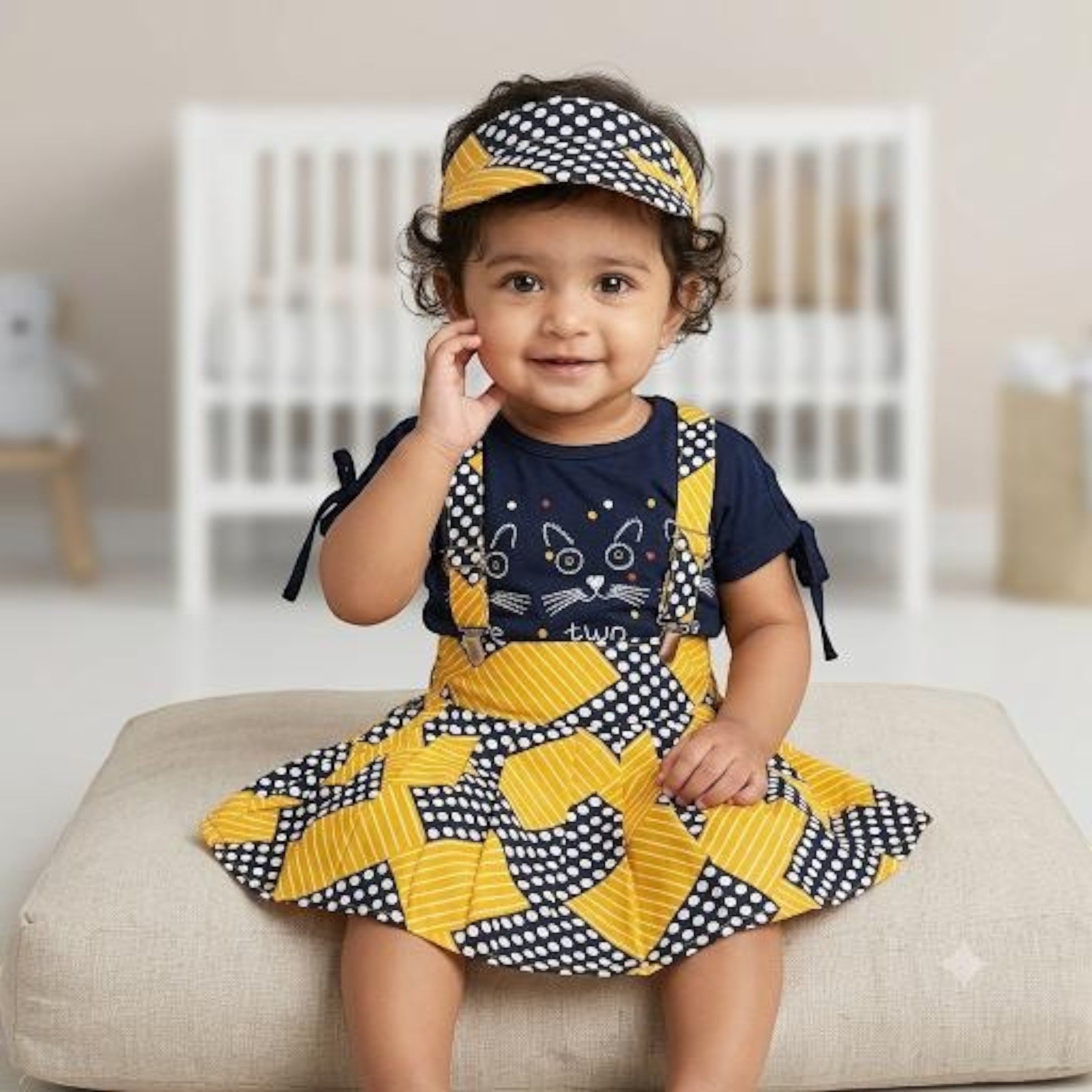 City Center baby girl wearing a navy blue cat-print top with a yellow and navy polka dot skirt and matching headband.