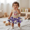 City Center baby girl wearing a beige graphic t-shirt and a purple floral flared skirt in a bright nursery setting.