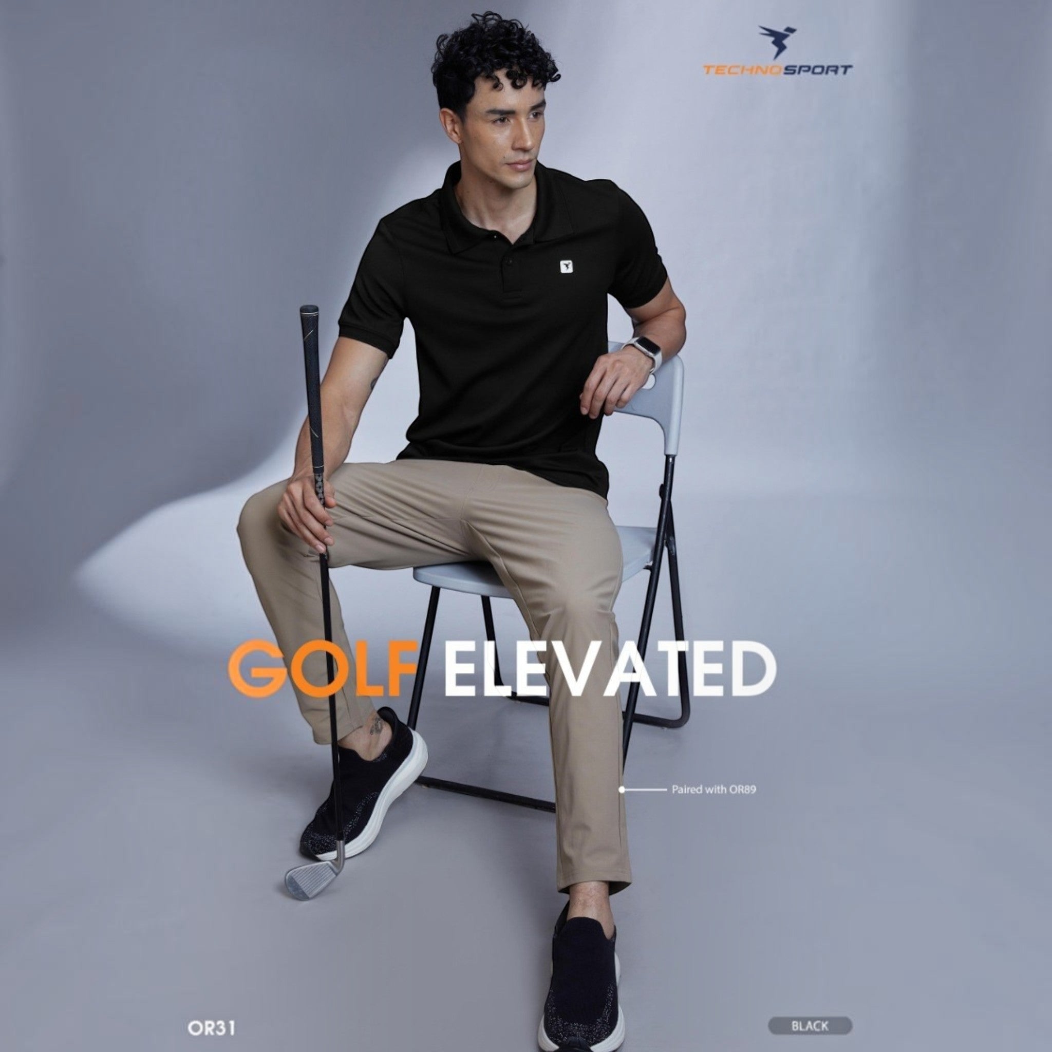 City Center OR31 black polo shirt worn by a male model sitting on a chair holding a golf club in a studio setting