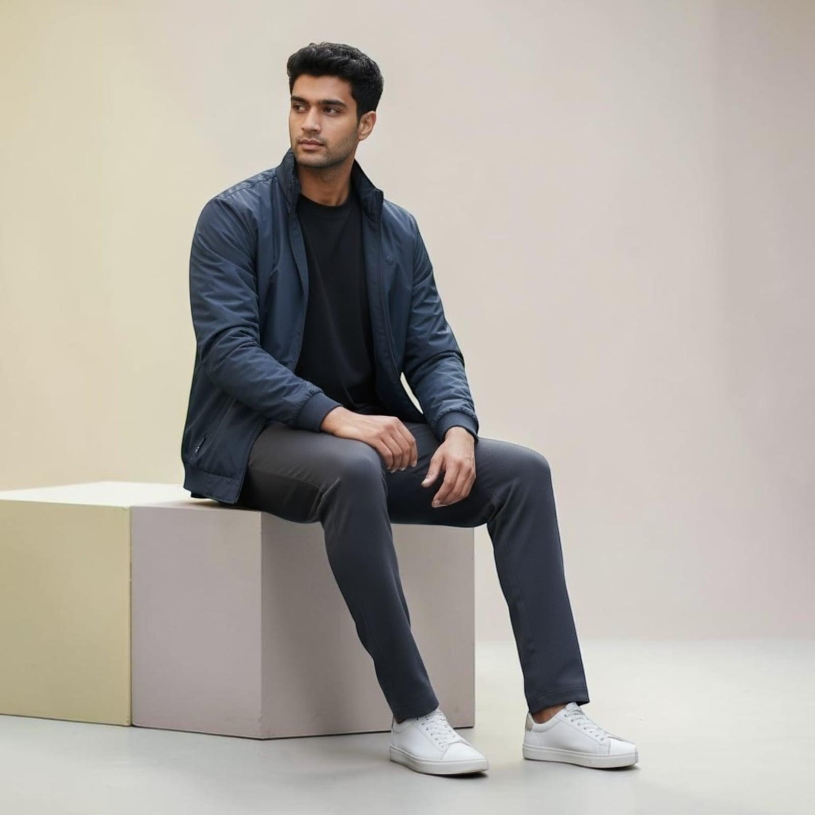 City Center men's navy blue zip-up jacket worn by a model sitting on a cube, paired with dark trousers and sneakers.