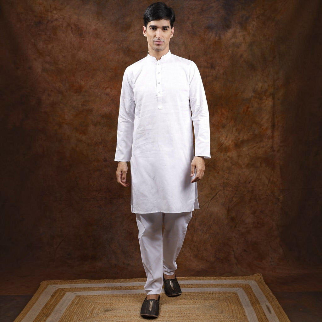 A man wearing a City Center white cotton kurta pyjama set, standing on a jute rug against a rustic brown backdrop.